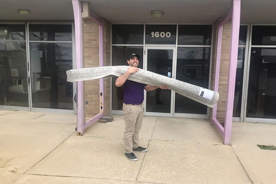 Man carrying a rolled-up area rug outside a building with purple columns and glass doors, ready for area rug cleaning services.