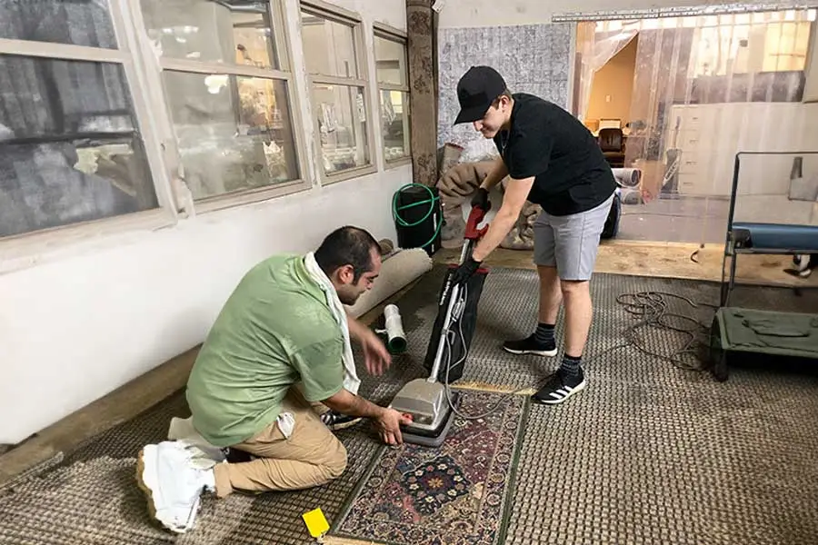Two men in a workshop cleaning an area rug, one kneeling and adjusting a rug cleaning machine while the other stands holding the machine's handle.