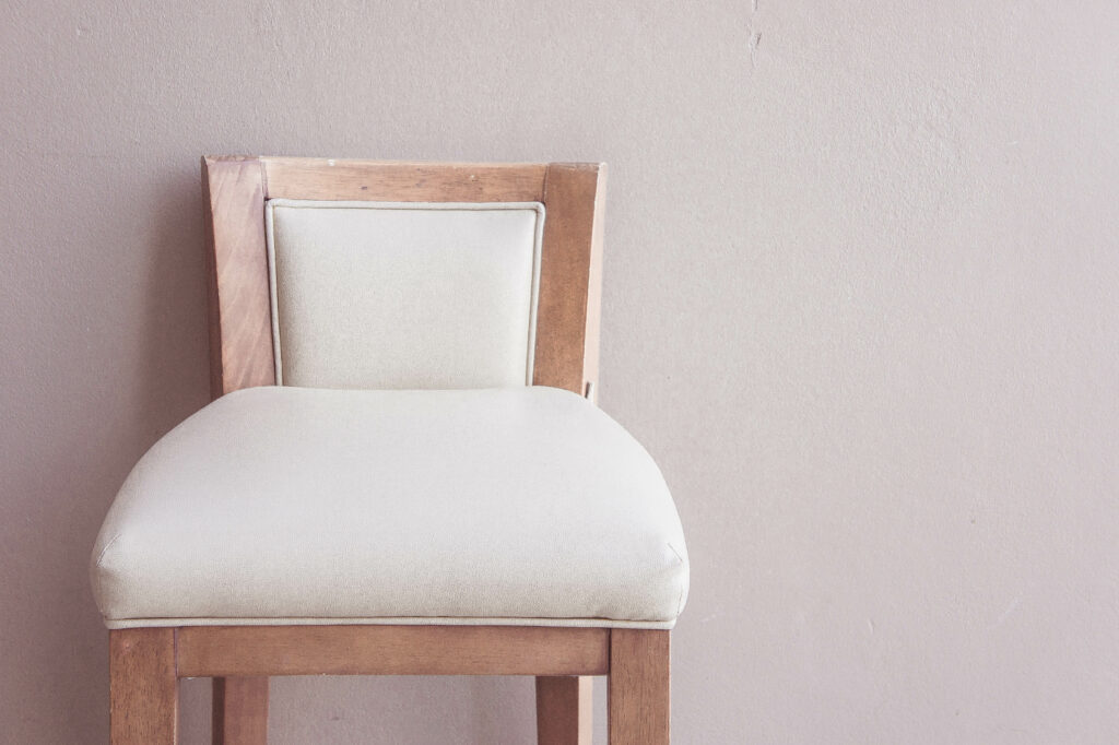 A wooden chair with light beige upholstery against a plain wall, illustrating a clean and simple design, and demonstrating the importance of regular upholstery cleaning to preserve its pristine appearance through professional upholstery cleaning techniques.