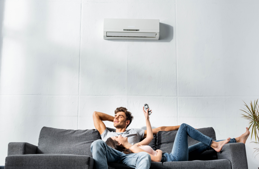 Couple relaxing on a sofa beneath a wall-mounted air conditioning unit, illustrating the comfort of clean indoor air and tips on how to get rid of mold in air ducts.