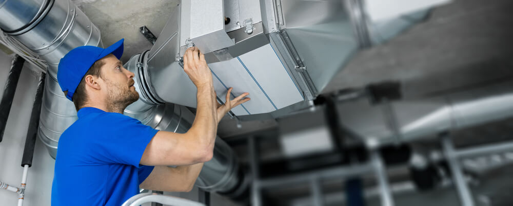 A technician in a blue uniform repairing a ventilation system, demonstrating the importance of clean air ducts. Discover what air duct cleaning do and the 5 benefits for your home.