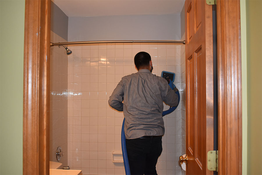 Worker from a tile and grout cleaning services company meticulously scrubs white wall tiles in a shower with a blue-handled brush, ensuring a sparkling clean finish, showcasing the results of professional tile and grout cleaning.